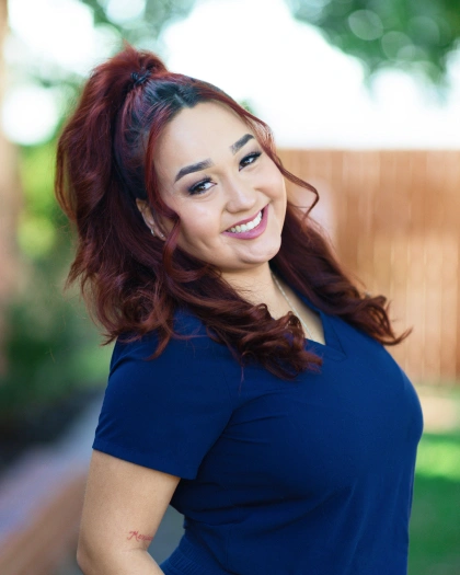 América Medina, Lab Technician Assistant at Warwick Dental, smiling in a navy scrub top, with a blurred outdoor background.