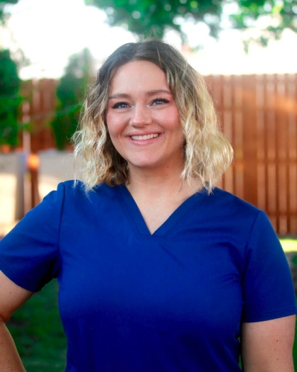 Courtney Carey, dental assistant at Warwick Dental, smiling in a blue scrubs top, outdoors with a wooden fence in the background, reflecting her commitment to patient care and creating smiles.