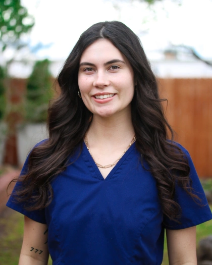 Hanna Smith, Front Office Insurance Claim Coordinator at Warwick Dental, smiling in a blue medical scrub, outdoors with greenery in the background.