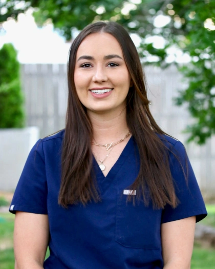 Jenna Perdue, registered nurse and dental assistant at Warwick Dental, smiling in a blue scrubs, outdoors with a blurred background of greenery.