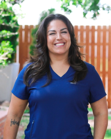 Maritza Spears, Lead Dental Assistant at Warwick Dental, smiling in a blue scrubs top, standing outdoors with a wooden fence and greenery in the background.