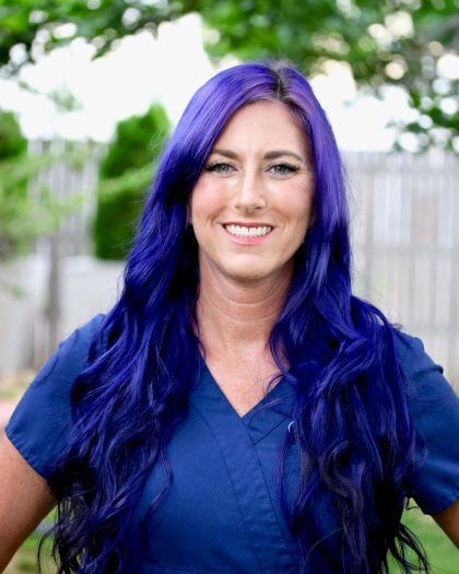 Sara Lyon, Registered Dental Hygienist at Warwick Dental, smiling with long purple hair in dental scrubs, set against a green outdoor background.
