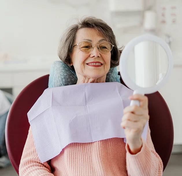 Elderly woman smiling while looking in a mirror at a dental clinic, wearing a dental bib, reflecting on her dental health after implant treatment.