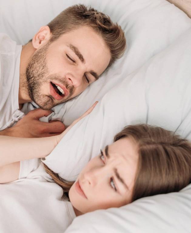 Man snoring in bed while woman looks concerned, illustrating sleep apnea symptoms and the impact on sleep health.